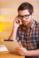 concentrado en el trabajo. un joven confiado hablando por teléfono móvil y mirando una tableta digital mientras se sienta en un café foto