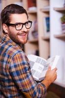 Handsome bookworm. Rear view of cheerful young man holding book and looking over shoulder while standing in front of the bookshelf photo