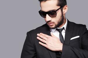 Used to look perfect. Handsome young man in sunglasses and formalwear touching his shoulder with hand while standing against grey background photo