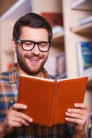 Reading his favorite book. Handsome young man reading book and smiling while standing in front of the bookshelf photo