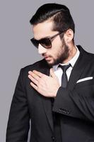 Style chaser. Handsome young man in sunglasses and formalwear touching his shoulder with hand while standing against grey background photo