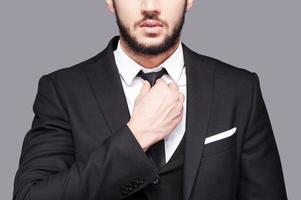 Corporate confidence meets exceptional style. Cropped image of fashionable young man adjusting his necktie while standing against grey background photo