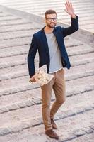 Hi there Full length of cheerful young man in smart casual wear holding bouquet of flowers and waving hand to someone while walking by the street photo