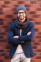 Confident in his perfect style. Handsome young man in smart casual wear keeping arms crossed and smiling while leaning at the brick wall photo