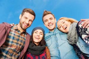 The best friends ever. Low angle view of four young happy people bonding to each other and smiling with blue sky as background photo