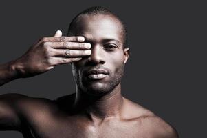 True masculinity. Portrait of young shirtless African man covering one eye with hand and looking at camera while standing against grey background photo