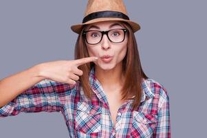 Kiss me Surprised young woman in glasses and funky hat pointing her lips and smiling while standing against grey background photo