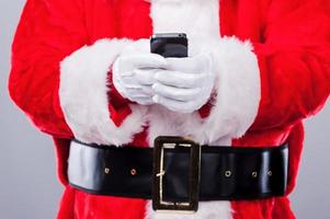 mensajes de texto de santa claus. primer plano de santa claus tradicional sosteniendo teléfono móvil mientras está de pie contra el fondo gris foto