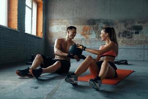 feliz pareja en forma haciendo ejercicio con balón medicinal mientras se sienta en colchonetas de ejercicio en el gimnasio foto