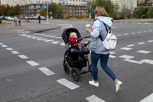una joven madre con un cochecito cruza la calle en un paso de peatones con una taza de café en las manos foto