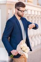 Esperándola. un joven apuesto con ropa informal elegante sosteniendo un ramo de flores y mirando su reloj mientras estaba parado en la calle foto