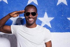 Yes sir yes Happy young African man in sunglasses holding hand near forehead and smiling while standing against American flag photo