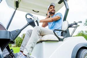 Estoy en camino al próximo hoyo, un joven apuesto y sonriente que conduce un carrito de golf y habla por teléfono móvil. foto