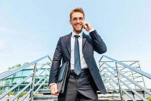 Businessman on the phone. Low angle view of confident young man in formalwear talking on the mobile phone and smiling while moving down by staircase photo