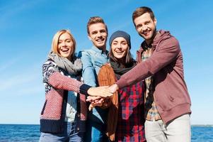 somos los mejores amigos. cuatro jóvenes felices uniéndose entre sí y tomándose de la mano mientras están de pie en la playa con el río en el fondo foto