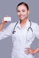 Helping every person who calls me. Confident female doctor in white uniform holding business card and looking at camera and smiling while standing against grey background photo