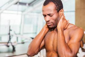 Feeling neckache after workout. Frustrated young African man touching his neck and expressing negativity while standing in gym photo