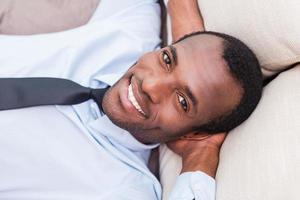 Taking time to for a minute break. Top view of handsome young African man in shirt and tie holding hands behind head and smiling while lying on the couch photo