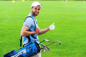 me encanta jugar al golf vista trasera de un joven golfista feliz que lleva una bolsa de golf con los conductores y mira por encima del hombro mientras está de pie en el campo de golf foto