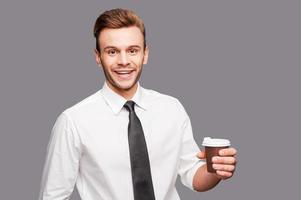 Take a break Cheerful young man holding a cup of coffee while standing against grey background photo