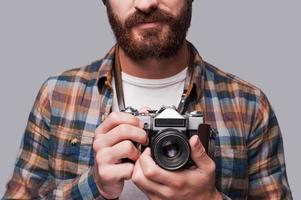 Smile to the camera Close-up of young bearded man holding old-fashioned camera while standing against grey background photo