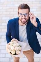 Well hello Top view of cheerful young man in smart casual wear holding bouquet of flowers and adjusting his eyeglasses while standing outdoors photo