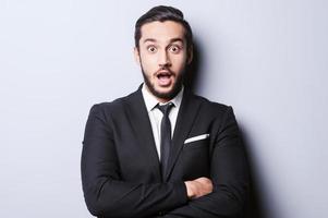 That is unbelievable Surprised young man in formalwear keeping mouth open and looking at camera while standing against grey background photo