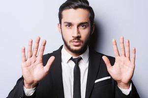 Stop it Serious young man in formalwear showing his palms and looking at camera while standing against grey background photo