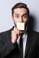 With humor to the deals. Portrait of surprised young man in formalwear covering his mouth with the sticky note while standing against grey background photo