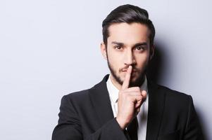 Keep my secret  Serious young man in formalwear holding finger on lips and looking at camera while standing against grey background photo