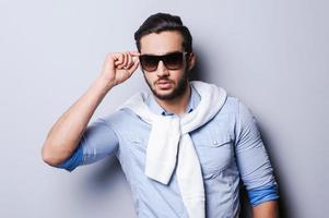 Style is my game. Handsome young man in blue shirt adjusting sunglasses while standing against grey background photo