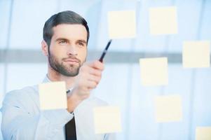 Thinking about solution. Thoughtful young businessman pointing adhesive notes attached to the glass photo