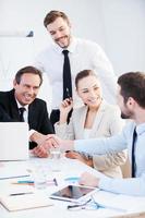 Congratulations Two confident businessmen handshaking and smiling while sitting at the table together with their colleagues photo