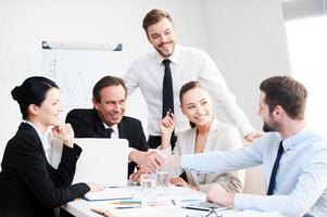 Welcome on board Group of confident business people in formalwear sitting at the table together and smiling while two men handshaking photo