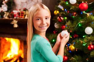 Preparing to Christmas. Cute little girl decorating Christmas Tree and smiling with fireplace in the background photo