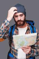 Lost in somewhere. Frustrated young bearded man carrying backpack and examining map while scratching head and standing against grey background photo