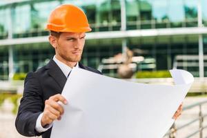 Architect examining blueprint. Handsome young man in hardhat examining blueprint while standing outdoors and against building structure photo