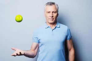 Healthy food for success. Confident senior man in casual throwing a green apple up and smiling while standing against grey background photo