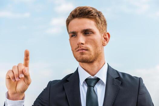 Working on transparent wipe board. Confident young man in formalwear touching a transparent wipe board while standing against sky background photo