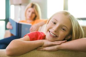 Enjoying time near mother. Happy little girl sitting on the couch and smiling while her mother reading book in the background photo