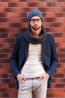 Confident handsome. Handsome young man in smart casual wear holding hands in pockets and looking at camera while standing against brick wall photo