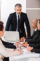 Confident and bossy. Business people in formalwear sitting at the table together while their boss standing and looking at them photo