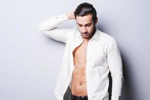 Real macho. Handsome young man in unbuttoned shirt holding hand on head while standing against grey background photo