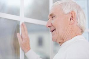 What a beautiful view Cheerful senior man keeping hand on window frame and looking through the window photo