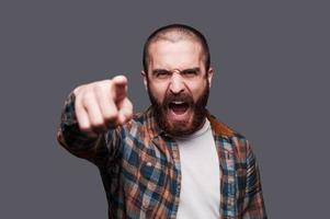 Hey you Furious young bearded man pointing you and shouting while standing against grey background photo