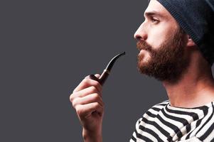 Just me and my pipe. Side view of confident young bearded man in striped clothing holding a smoking pipe and looking away while standing against grey background photo