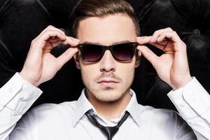 Confident handsome. Handsome young man in shirt and tie adjusting his sunglasses while standing against black background photo