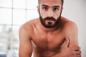 Bearded handsome. Confident young bearded and shirtless man looking at camera while sitting indoors photo