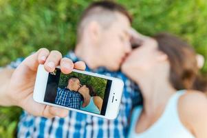 Capturing a moment. Top view of happy young loving couple making selfie with smart phone while lying on the grass and kissing photo