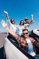 Fun travel. Group of young happy people enjoying road trip in their white convertible and raising their arms up photo